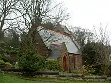 A small church seen from the southwest with a south porch. At the far end a saddleback-roofed tower is just visible. Around the church are trees and bushes and in the foreground is a wall