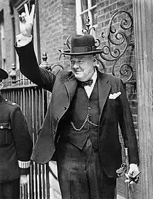 U.K. Prime Minister Winston Churchill in a black lounge suit with homburg hat and walking stick on Downing Street, giving his famous 'V' sign during World War II.