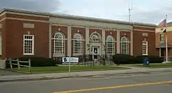 A one-story brick building with elaborate classically-inspired stone detailing, seen from across a street. There are five round-arched windows grouped in the center and two rectangular ones nearer the ends. At the right is an American flag flying from a pole and a mailbox.