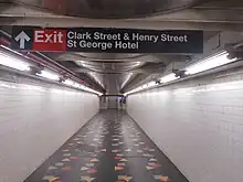 A passageway above the platforms, leading to the elevators. The passageway has black floors with multicolored shapes, which are part of an artwork. The walls are clad with white tiles. There is a sign on the ceiling, directing passengers to the station's exit in the St. George Hotel.