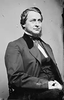 A pale-eyed man sits in a chair, looking to the right. He is wearing a dark formal suit and has a Donegal beard.