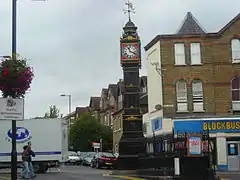 The Clock Tower in South Norwood. It stands on a traffic island. It is a black cast-iron tower, with gilt decoration. The clock face surround is red, with the face being white, with black hands and figures. At the top of the tower is a wind-vane, featuring an arrow and the cardinal compass directions.