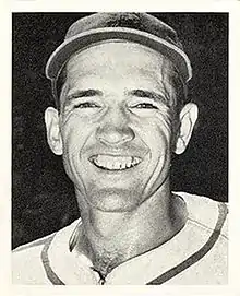 A smiling man in a white baseball uniform with dark trim going around the neck and a dark cap.