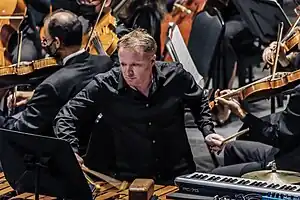 Colin Currie, wearing black t-shirt and black trousers, holds two drumsticks towards the camera, classical musicians are pictured behind him.