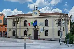 Wide yellow building has a clock at the top, and flies the EU and Italian flags.