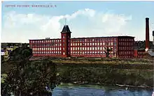 A tree canopy at bottom left obscures a river running left to right, behind which the river bank rises several storeys. Set back from the bank, and viewed at a slight angle, is a four-storey red building with long rows of windows. The middle of the building has a five-storey tower capped by a square pyramidal roof truncated at the top. At the far right and separated from the building is a chimney, about seven storeys tall.