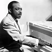 Posed photo of a man with short hair and a short mustache, wearing a suit, seated at a grand piano with his hands on the keys