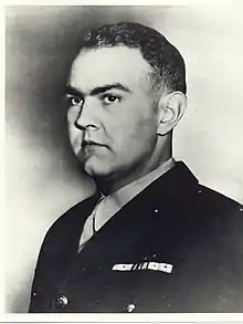 Head of a stern-faced young white man with dark hair and full cheeks wearing a dark jacket with ribbon bars of the left breast.