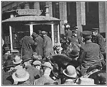 A crowd of men swarm around a streetcar and mounted police officers