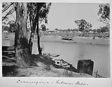 In the foreground a large tree on a riverbank; across the river is a mission station with numerous huts