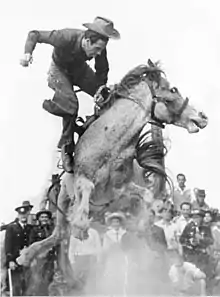 A man dressed in cowboy-style is in mid-air above the back of a horse bucking in a cloud of dust