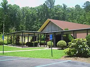 A single-story building with a high peaked reddish-brown shingled roof, a large triangular paned window divided by white strips, yellowish eaves, and some brown brick facades. In front of it are walkways surrounded by an open poles and a lighted horizontal covering. A well-kept grass lawn is in front of that, with a number of trimmed green shrubs and bushes all around, one of which on the right has bluish blossoms. In front of the lawn, an asphalt parking area, with spots marked in blue and blue signs indicating handicapped parking. Behind the building, an open slope with some telephone wires and then behind that, a continuous stand of tall deciduous trees.