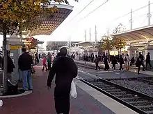 Numerous passengers walking and waiting along the tracks at the side platforms of the station with a  train and arena at rear.