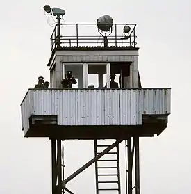 On top of a tubular steel support structure with a ladder there is a small cabin with windows to all sides, surrounded by a balcony on which stand two men in uniform, looking at the observer through binoculars; a third man is inside the cabin. There are searchlights on the tower's flat roof, and a railing around the roof's outer edge.