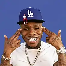 Cover art depicting DaBaby smiling and pointing finger guns at his head.  He wears a baseball hat and a smaller animated version of himself is sitting on the brim.