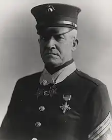 Head and shoulders of a white man wearing a cap and a jacket with stripes on the upper sleeves, one medal pinned to the left breast, and two identical star–shaped medals hanging from ribbons around his neck.