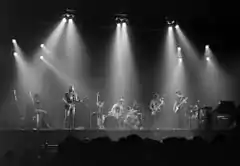 A monochrome image of members of the band. The photograph is taken from a distance, and is bisected horizontally by the forward edge of the stage. Each band member and his equipment is illuminated from above by bright spotlights, also visible. A long-haired man holds a guitar and sings into a microphone on the left of the image. Central, another man is seated behind a large drumkit. Two men on the right of the image hold a saxophone or a bass guitar and appear to be looking in each other's general direction. In the foreground, silhouetted, are the heads of the audience.