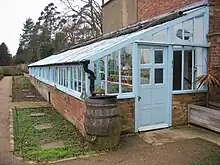 Lean-to greenhouses against a brick wall, with an entrance door and water butt at the nearest end and trees in the background