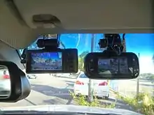 A pair of black devices with small monitors stuck with suckers to the inside of a car windshield with blue tint at the top, seen from inside the vehicle