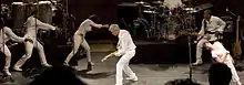 Performers on the tour in a semi-circle bowing and gesturing toward Byrne as he plays guitar