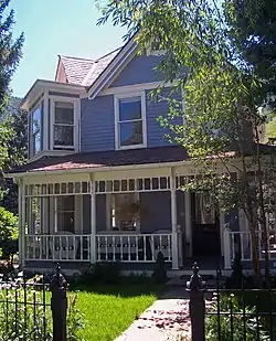 A light blue wooden house with pointed roof and a covered white-trimmed porch. There is a square projecting section on the left corner. The view is slightly obscured by trees; at the edges a distant ridgeline can be seen. In the foreground, at the bottom, is a pink stone walk and black iron fence