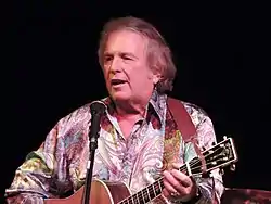 A grey-haired man in a brightly-patterned shirt playing a guitar and singing into a microphone