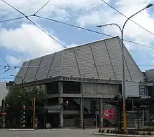 two stry concrete builign with metal angled roof. feels squat and is a brutalist style of architecture. bus trolley wires in the foreground over the street