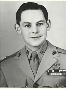 Head and shoulders of a young white man with neatly combed dark hair wearing a light-colored military jacket with three rows of ribbon bars and a parachute pin on the left breast.