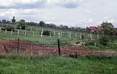 A hillside with multiple barbed-wire fences running parallel to each other, with fruit trees, a barn and a watchtower in the background.