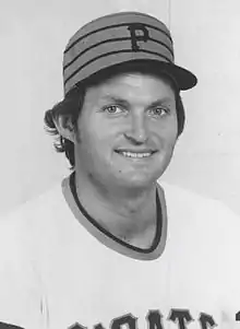 A man smiles for the camera while wearing a white baseball jersey and a cap with three stripes going around and a "P" on the front.