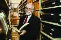 A photograph taken in November 2010, of then Maryland State Archivist and Commissioner of Land Patents, Dr. Edward C. Papenfuse, holding a book, possibly which are bounded records, while standing with a smile on his face, in a row of shelved stacks of other bounded books, in the archive.