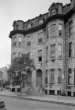 End section of a four-story stone terraced building. Two adjacent doors are at center, each with a short flight of stairs leading up from the street to the first level. Either side of the doors, at left and right, the building has two rounded tower-like sections, each with nine large sash windows (three on each level). Four more large windows are at center, above the doors. The uppermost (attic) level has four smaller windows protruding from the roof with architectural feature. At street level, grilled skylights illuminate basement rooms.