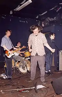 Three people are standing on a stage, while one is sitting and playing the drums. One person is playing the guitar.