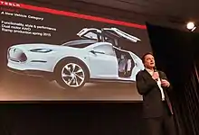 Musk speaking with a microphone, a projected image of a Tesla is behind him