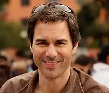 A caucasian male with dark hair, wearing sunglasses on the top of his head, with a brown suede jacket, smiles