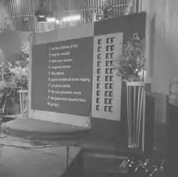 Black and white photograph of the scoreboard in 1958; the running order numbers and song titles of the competing entries are printed on the left-hand side of the scoreboard, and rotating numbers on the right-hand side show the allocation of points to each song as each country's jury is called, and a total of all points received; song titles are sorted by order of appearance, with the first song to be performed appearing at the top of the scoreboard.