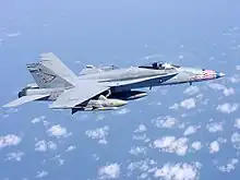 Overall-gray jet fighter, with red, blue and white-tipped nose, is overflying sea and scattered white clouds down below. The aircraft is carrying streamlined external fuel tanks and missiles under its wings, and is heading right.