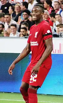 Isaac Mabaya, playing football for Liverpool, wearing their home shirt, he is standing in front of advertising boards with fans in the background