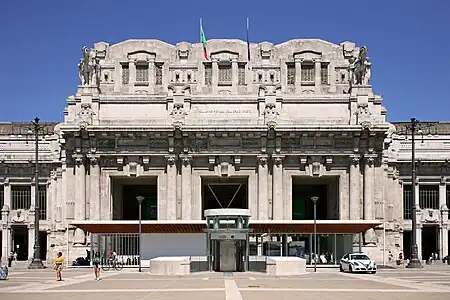 An ornate and extensively detailed gray stone classical portico with four sets of twinned columns at the top of which the Italian and European Union flags hang limply; above is clear blue sky. In front is a more modern one-story pavilion with an elevator in front of it inside a circular wall. Some people can be seen walking around.