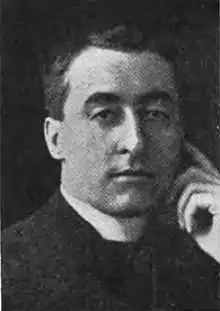 Black and white photograph of a man wearing a Roman collar from the mid-chest up. He faces the camera and touches his index finger to his temple.