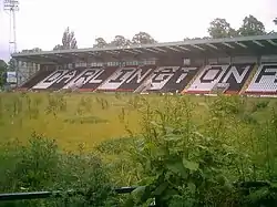 A grandstand whose coloured seats spell out the word Darlington. A floodlight pylon stands at one end. The stand faces a grassed area overgrown with weeds.