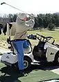 A golfer with a leg amputation uses an adaptive golf cart that has a chest strap to help him maintain his balance while standing on one leg.