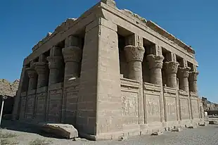 Small stone building surrounded by half-walls and columns with elaborate capitals.