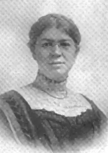 A white woman wearing glasses and a dress with a high lace collar; she has dark hair parted center and braided over the crown