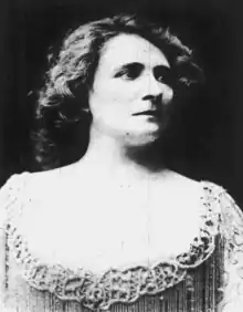 A white woman with wavy hair, wearing a dress with a wide, low, lace-trimmed neckline