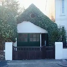 An extremely small workshop adjoining a house and standing behind a wooden gate with white gateposts. It has green double doors with an open door to their right.  Above this is a white-painted wall which is heavily draped with ivy, although a circular louvre is clearly visible through this.  The roof is steep and pointed.