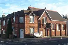 A dark red brick hall on a corner site, with adjoining houses in an identical style. The hall, partly obscured by a traffic light, has a pointed-arched entrance with a wooden door below a rectangular window frame with three lancets.  To the right of the door, there are three plain windows between brick buttresses.  Above the entrance is a steep roof perpendicular to the main roof.