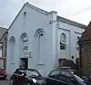 Three-quarter view of a pale blue building with a plain exterior and a large pediment, on a crowded site between two flint structures. The façade has three tall, triple-recessed blank arches, the centre of which is shortened to accommodate an entrance door. A round opening sits just below the pediment. The side has round-arched windows.