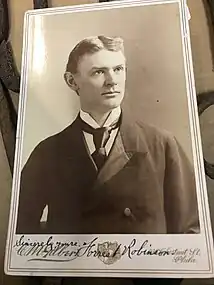 This is a black-and-white photograph of a slender young man with a handwritten signature identifying him as Forrest Robinson at the bottom.  The photo captures the man from the chest up with his shoulders facing the front and his head turned slightly to the right against a gray background.  He has short, apparently blond or light brown hair parted in the middle and combed back above the ears and light colored eyes.  He is clean shaven and has a calm expression with lips closed and eyes directed to the right of the viewer. He is wearing a dark, double-breasted frock coat made of wool with tulip-shaped lapels.  A satin or silk lining can be seen on the inner part of the lapel.  A crisp, white shirt with a high collar and folded corner wings and a large black tie made of a satin-like material are visible in the short cleavage between the lapels.  The coat has visible wrinkles.  The photograph is glued to a cardboard backing.  The name of the studio, C. M. Galbert, is written in cursive font on the left side and the address of the studio is written on the right side in a smaller cursive font in two lines:  illegible number Cherry St., Phila.  There is a heraldic emblem centered beneath the photograph.  The signature appears in black ink just studio name on the far left with the dedication, Sincerely yours, and the name Forrest Robinson is written in a larger cursive script across the heraldic emblem and the first part of the address.  Two thin lines create a frame around the photo and the text.