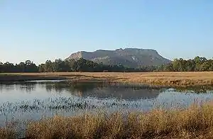 Daytime view of a small marshy lake surrounded by dry brush in winter. Behind the lake in the far-middle distance, a large stand of trees interposes itself in front of a lone massif in the far distance. The sun is out of view to the left.